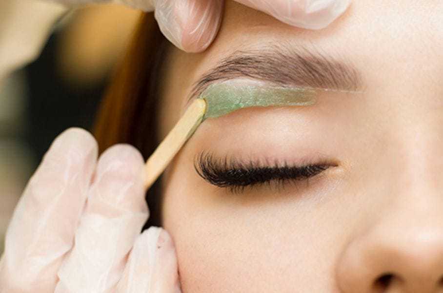 Eyebrow waxing process with green wax applied on closed eye.